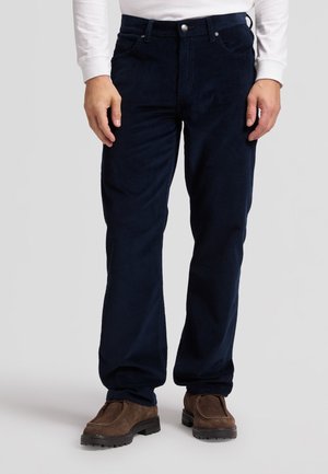 Homme portant un pantalon en velours côtelé bleu foncé, des chaussures en daim marron et une chemise blanche à manches longues sur un fond clair uni.