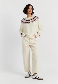 Maglione crema con motivo a diamante bordeaux attorno al colletto. Abbinato a pantaloni larghi crema e sneakers bianche con accenti grigi.