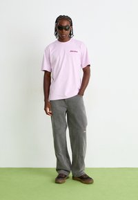 Rosa T-skjorte med kort erme og "Dickies"-logo, grå baggy bukser, og mørke solbriller, kombinert med brune teksturerte sko på en grønn overflate.