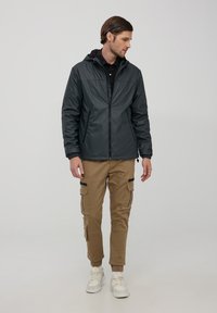 Giacca leggera grigia con cappuccio, zip frontale, polsini elastici, abbinata a pantaloni cargo beige con tasche laterali e cerniere. Scarpe da ginnastica bianche.