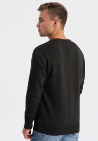 Felpa nera con collo e polsini a costine, caratterizzata da un tessuto strutturato. Il modello la indossa con jeans azzurri, vista da dietro.