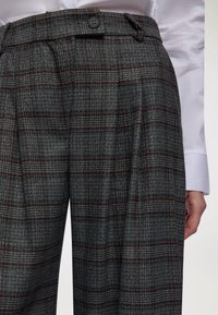 Pantaloni a quadri grigi con accenti bordeaux indossati con una camicia bianca, che mostrano la vita, il bottone e la mano laterale di chi li indossa.