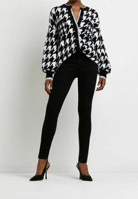 Svartvit houndstooth-kardigan med knäppning fram, vida ärmar och kontrasterande grå detaljer, kombinerad med smala svarta byxor och klackar.