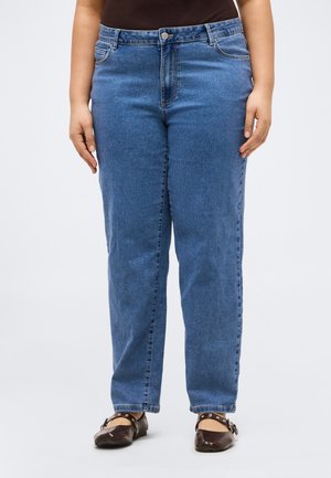 Mujer con jeans azules de talle alto y pierna recta, con botones y cremallera, combinados con zapatos planos marrón oscuro con correas en el tobillo.