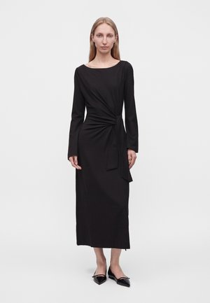 Femme aux longs cheveux blonds portant une robe midi noire à manches longues avec un détail de nœud latéral et des chaussures plates noires pointues, debout sur fond blanc.