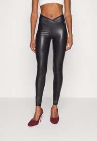 Kobieta w czarnych, wysokich legginsach z ekoskóry i bordowych, teksturowanych szpilkach, stojąca przy gładkiej ścianie.