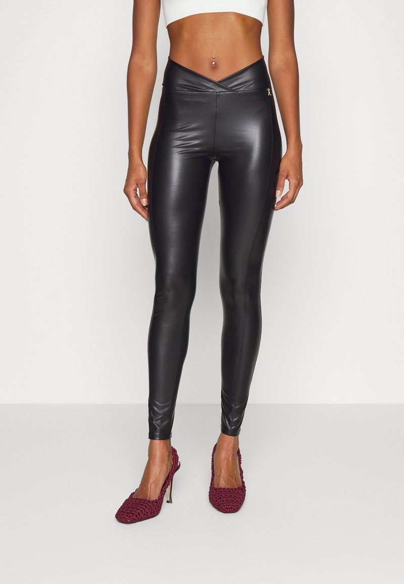 Kobieta w czarnych, wysokich legginsach z ekoskóry i bordowych, teksturowanych szpilkach, stojąca przy gładkiej ścianie.