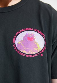 Fekete pamut póló, amelyen egy béka grafikája látható, rózsaszín és lila akcentusokkal egy kör alakú dizájnban. Szöveg veszi körül a képet.