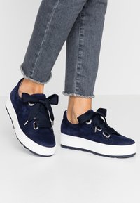 Sneakers in suede blu navy con lacci in nastro oversized e occhielli in metallo. Presentano una suola piatta bianca e testurizzata. Visibili jeans con orlo sfrangiato.