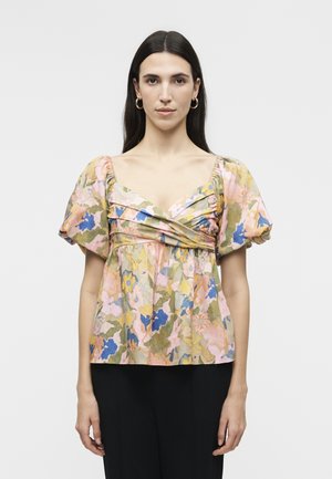 Γυναίκα με ίσια σκούρα μαλλιά, που φορά χρυσούς κρίκους για σκουλαρίκια και μια φλοράλ μπλούζα με φουσκωτά μανίκια και λαιμόκοψη που διασταυρώνεται, στέκεται μπροστά σε λευκό φόντο.