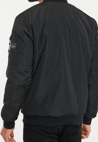 Giacca bomber nera con una texture liscia e lucida. Presenta polsini e fascia in vita a costine, e una tasca con zip sulla manica sinistra.