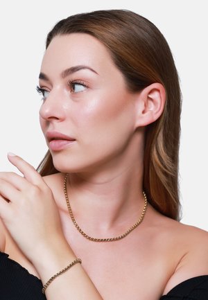 Collier en chaine dorée et bracelet portés sur la peau nue. Le collier présente des maillons ronds ; les deux accessoires exhibent une finition lisse et brillante.