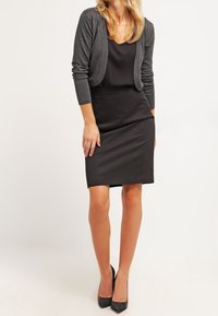 Schwarzes ärmelloses Kleid mit figurbetonter Silhouette, kombiniert mit einer dunkelgrauen Strickjacke. Das Outfit wird durch schwarze High Heels ergänzt.