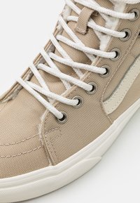 Beige høyhælte sneakers laget av teksturert lerret, med hvite snørebånd, metallisk øyelets, og en gummisåle med kontraststriper.