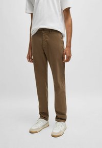 Bruine chino's van katoen, met een slim fit, zijzakken en een knoopsluiting, gecombineerd met witte sneakers op een effen achtergrond.
