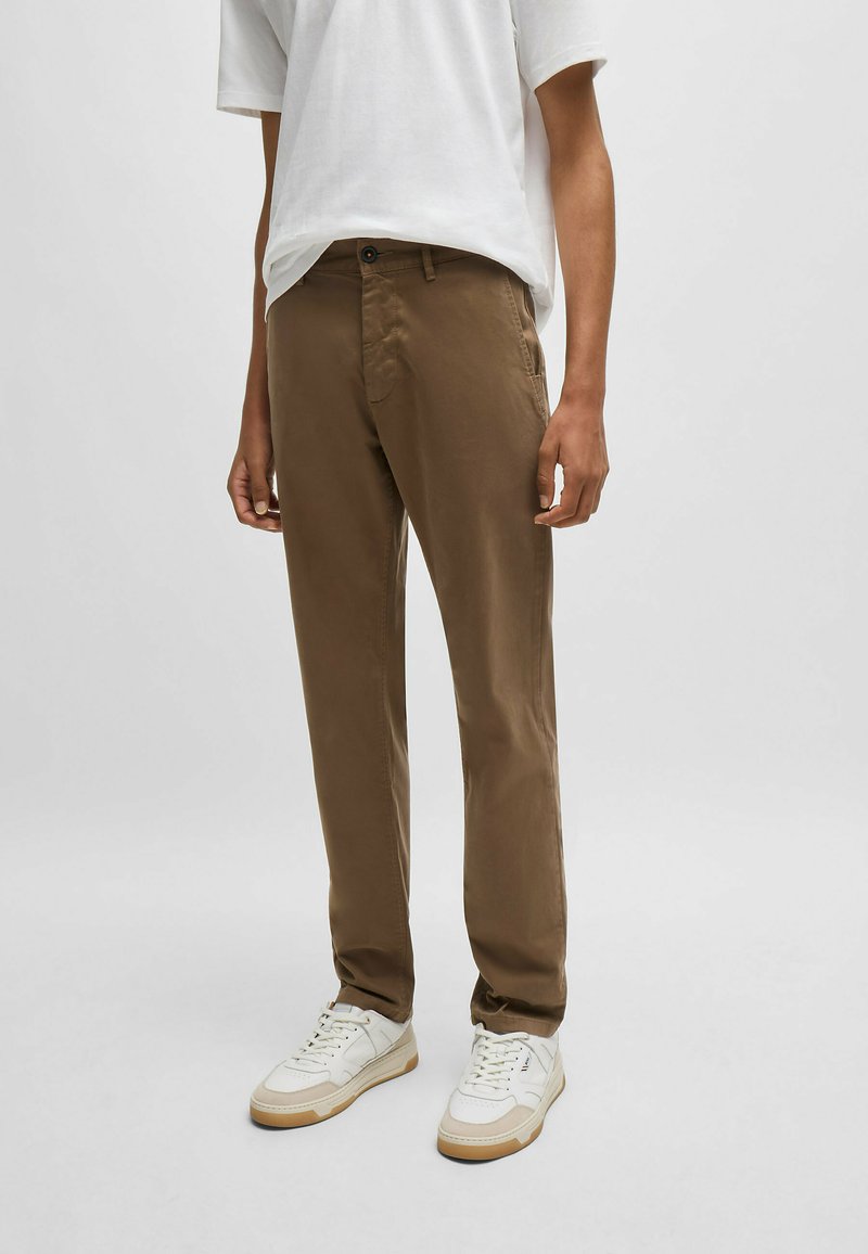 Bruine chino's van katoen, met een slim fit, zijzakken en een knoopsluiting, gecombineerd met witte sneakers op een effen achtergrond.