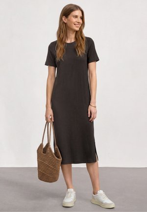 Femme portant une robe midi noire à manches courtes et des baskets blanches, tenant un sac fourre-tout tressé marron, debout devant un fond uni.