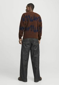 Brauner Strickpullover mit blauen Wellenmustern, kombiniert mit dunkelgrauen weit geschnittenen Jeans. Der Pullover hat gerippte Bündchen und einen gerippten Saum.