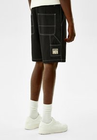 Musta cargo-shorts useita taskuja, valkoista ompelua ja merkkilappu. Yhdistetty valkoisten sneakerien ja ribattujen valkoisten sukkien kanssa.