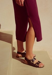 Sandales en cuir marron avec deux grandes boucles et semelles en liège, portées avec une robe prune mi-longue à fente latérale, posées sur des marches.