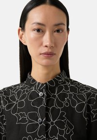 Black blouse with white floral outline pattern. Features a collared neck and two black buttons, made from a lightweight, textured fabric.