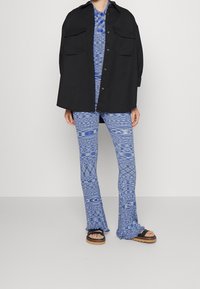 Chemise-veste noire surdimensionnée avec deux poches avant, associée à un pantalon évasé bleu à motifs et des sandales à plateforme noires.