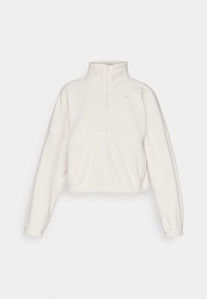 Pull en polaire, couleur crème clair, col zippé, coupe ample, poignets et ourlet élastiques, avec un petit logo en haut à gauche.