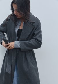 Grå ullblandningskappa med slagkrage, bälte i midjan och stora knappar. Modellen justerar en smartphone i fickan. Ljus bakgrund.