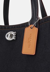 Sac à main en tissu noir avec coutures orange, poignées en cuir noir, fermoir à tourniquet argenté et étiquette en cuir marron clair de la marque Coach sur une chaîne à billes.