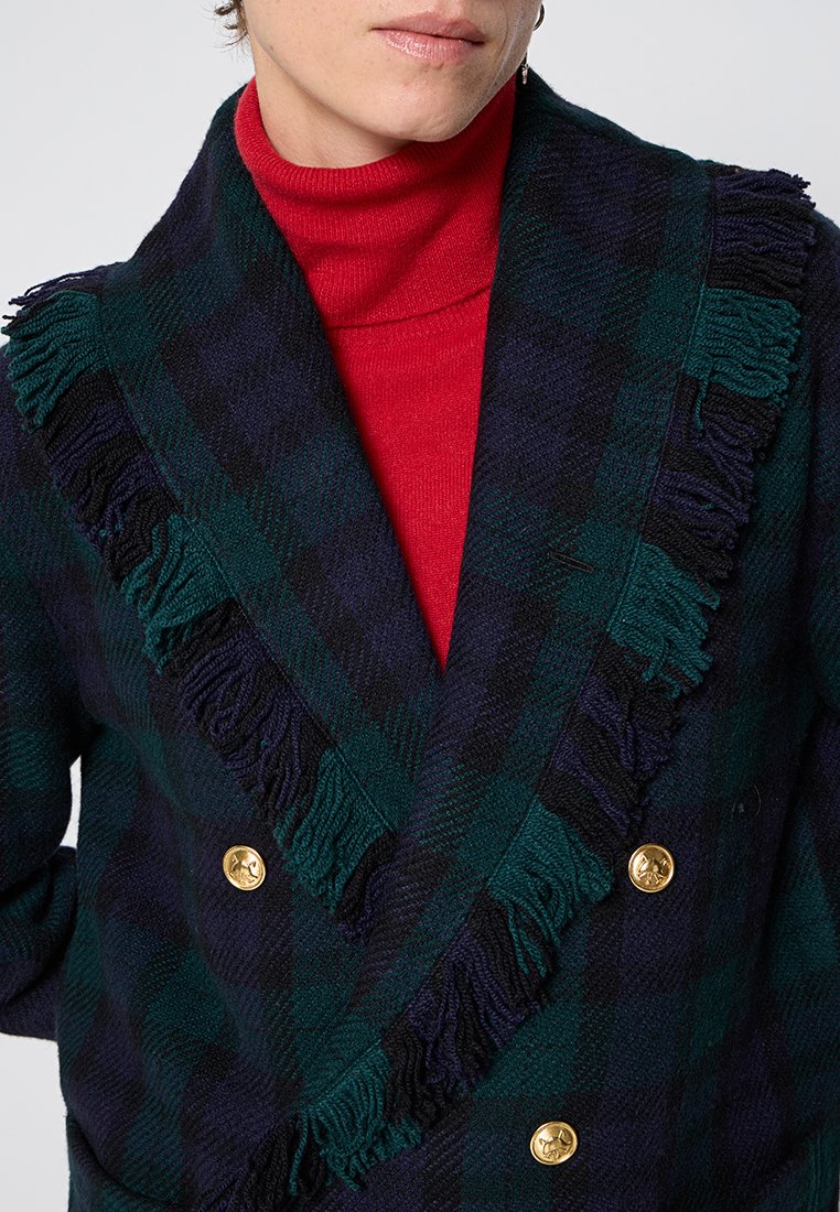 Osoba ubrana w ciemnozieloną i granatową w kratę frędzlowaną kurtkę z złotymi guzikami, noszącą jasnoczerwoną bluzę z wysokim kołnierzem, skróconą na wysokości podbródka.