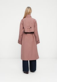 Femme aux longs cheveux blonds se tient de dos, portant un trench-coat rose poussiéreux sur un pantalon large noir et des chaussures marron.