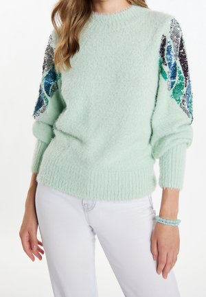 Kvinde iført en lysegrøn fuzzy sweater med flerfarvede palietdesigns på ærmerne og hvide bukser, tilbehør med et armbånd.