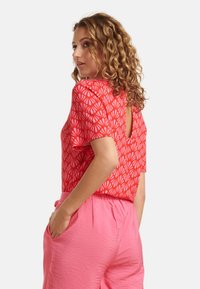 Rotes Kurzarm-Oberteil mit weißem abstraktem Muster und einem verlängerten Rückenteil, kombiniert mit rosa strukturierten Hosen und einer Taschenoptik.