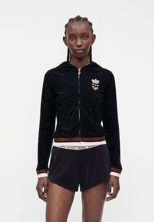 Femme aux cheveux tressés, portant un sweat à capuche noir zippé avec des bordures roses et marron et un short noir avec une ceinture blanche portant l'inscription « Juicy Couture ».