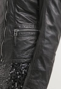 Giubbotto di pelle nera con una texture morbida, chiusura a zip diagonale e dettaglio della tasca laterale con zip. Parzialmente mostrato su uno sfondo di tessuto a paillettes.