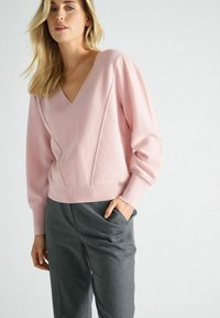 Pull over rose avec décolleté en V et manches longues bouffantes, présentant des détails de couture diagonale. Associé à un pantalon gris sur mesure. Texture douce.