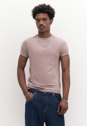 Göndör hajú fiatal férfi világos mályvaszínű pólóban, sötétkék farmerben, ezüst színű lánc nyaklánccal és karkötőkkel, fehér háttér előtt áll.