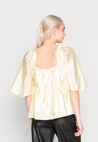 Top peplum amarelo em satin com mangas curtas e onduladas, cintura ajustada e costas abertas com detalhe de laço. Calças de couro pretas visíveis abaixo.