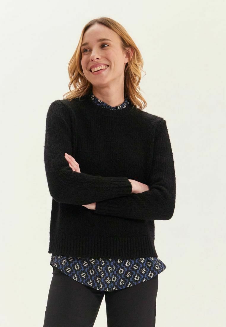 Pull en tricot noir avec une finition texturée, doté d'un col rond et superposé sur une chemise à motifs bleus avec un design floral.