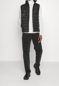 Musta toppaliivi, jossa korkea kaulus, kiiltävä pinta ja vaakasuorat tikkauskohdat. Päällä valkoinen pooloneule, yhdistetty tummiin housuihin ja mustiin kenkiin.