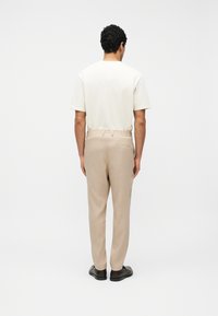 Άνδρας όρθιος με πλάτη, φοράει λευκό πουκάμισο με κοντά μανίκια, μπεζ παντελόνι με ελαστική μέση και μαύρα επίσημα παπούτσια σε λευκό φόντο.