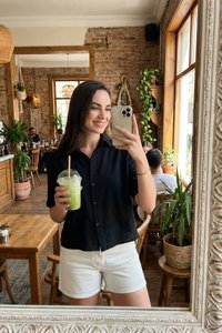 Ung kvinde smiler, holder en grøn smoothie og en smartphone, tager spejlselfie i en hyggelig café med planter og møbler af træ.