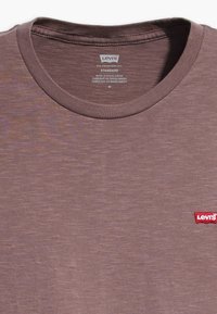 Braunes Baumwoll-T-Shirt mit einem gerippten Rundhalsausschnitt und subtiler Textur. Enthält ein kleines rotes Logo auf der linken Brust. Größe M.