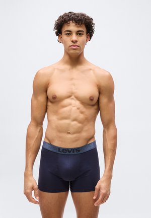 Ung mand med krøllet hår står med fronten mod kameraet, uden trøje, iført mørkeblå Levi's boxer shorts mod en ensartet lys baggrund.