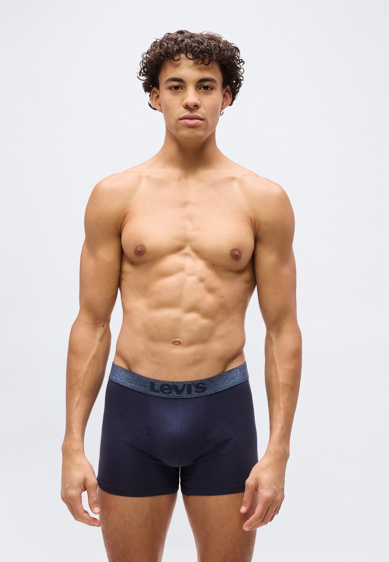 Jeune homme aux cheveux bouclés se tenant face à l'avant, torse nu, portant un boxer Levi's bleu foncé sur un fond clair uni.