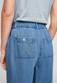 Back view of person wearing light blue elastic waist denim pants with two buttoned back pockets and a light grey tucked-in shirt.