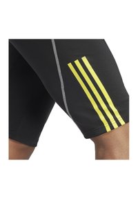 Zwarte sportshorts van rekbaar materiaal, met drie gele strepen aan de zijkant en grijze stiksels langs de naden.