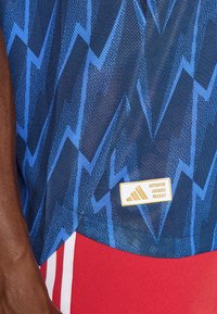 Marine sports top med blå lynmønstre, fremstillet af mesh-materiale. Har et guldlogo-patch mærket "AUTENTISK LICENSERET PRODUKT."
