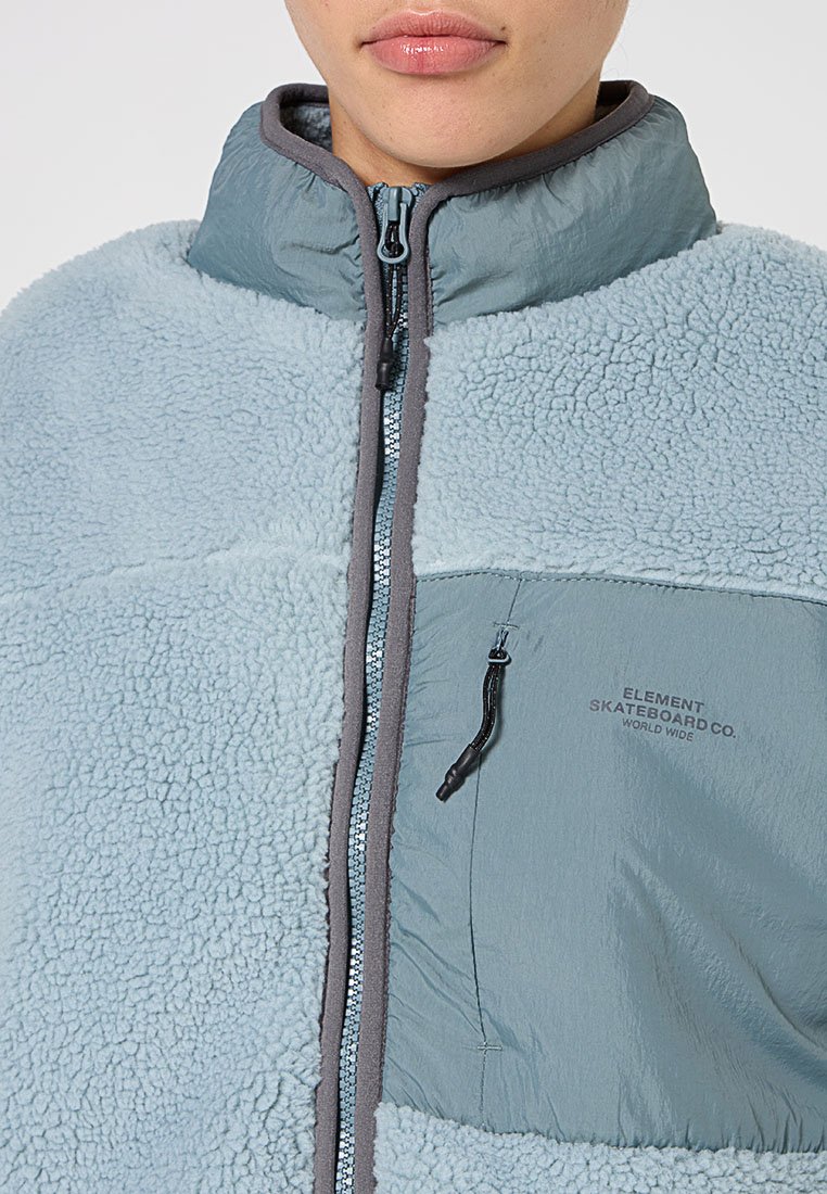 Helleblaue Kunstfelljacke mit Stehkragen. Merkmale einen Reißverschluss, eine Brusttasche und kontrastierenden grauen Besatz. Logos sind auf der Tasche angebracht.