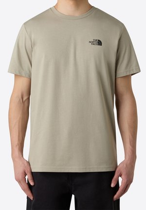 Homme portant un T-shirt beige à manches courtes et col rond avec un petit logo noir The North Face sur le côté gauche de la poitrine, assorti à un pantalon noir.